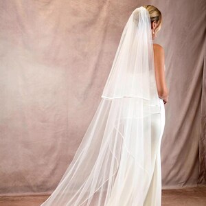May include: A long, two-tier white bridal veil made of sheer tulle fabric with a finished edge, worn over a floor-length white gown. The veil features a shorter top layer and a significantly longer, flowing train that extends across the floor.