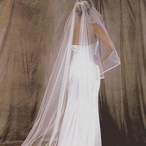 May include: A long, sheer white wedding veil featuring a delicate satin ribbon trim along the edges. The veil is attached to a hair accessory at the crown and cascades down the back, extending into a floor-length train over a white gown.