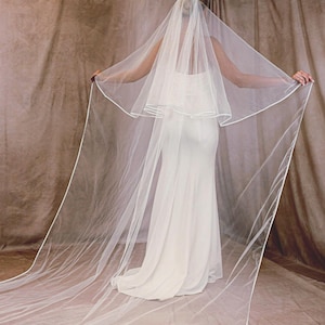 May include: A bride in a white wedding dress stands with arms outstretched, showcasing a long, sheer, white veil with a delicate white trim. The veil cascades down, creating a flowing effect. The background is a neutral brown.