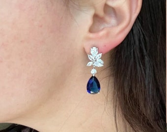 May include: Elegant drop earrings featuring a cluster of clear, sparkling stones at the top, with a single, large, pear-shaped, deep blue gemstone dangling below. The earrings are set in a silver-toned metal and are worn on an ear.