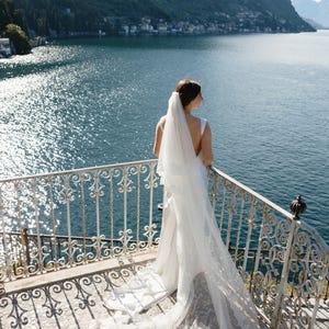 K&ouml;nnte beinhalten: Eine Braut in einem wei&szlig;en Hochzeitskleid und Schleier steht auf einem Balkon mit Blick auf einen See und Berge. Das Kleid hat eine lange Schleppe und der Schleier ist mit Spitze verziert. Der Balkon hat ein dekoratives schmiedeeisernes Gel&auml;nder.