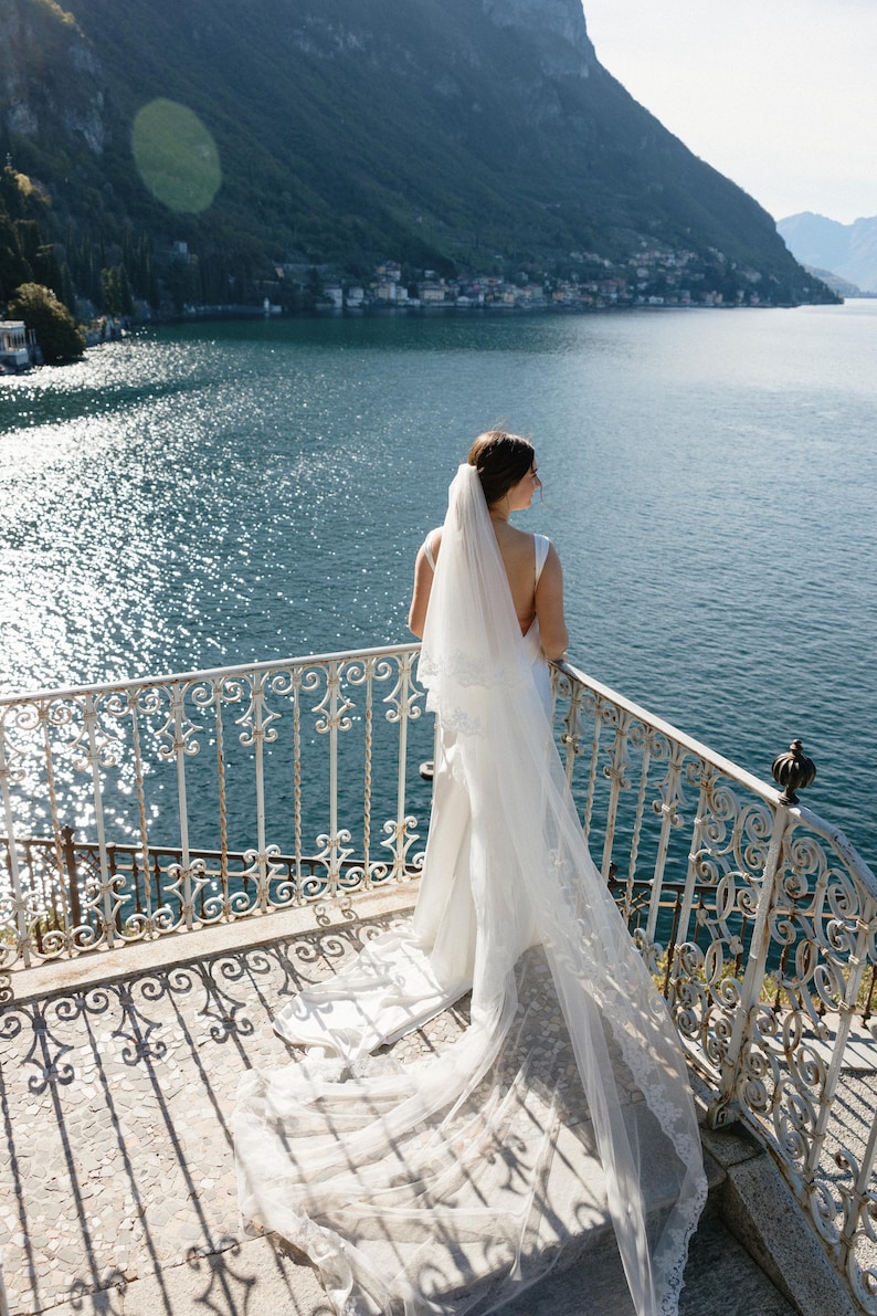 K&ouml;nnte beinhalten: Eine Braut in einem wei&szlig;en Hochzeitskleid und Schleier steht auf einem Balkon mit Blick auf einen See und Berge. Das Kleid hat eine lange Schleppe und der Schleier ist mit Spitze verziert. Der Balkon hat ein dekoratives schmiedeeisernes Gel&auml;nder.