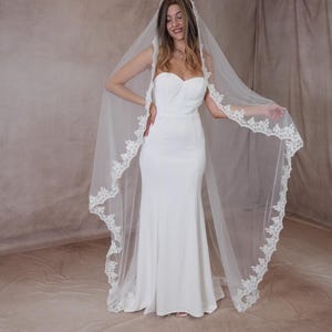 May include: A white bridal gown with a strapless design and a flowing skirt. A sheer, white veil with intricate lace edging drapes down the back. The bride is smiling, displaying the elegant wedding attire.