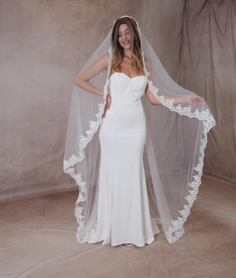 May include: A white bridal gown with a strapless design and a flowing skirt. A sheer, white veil with intricate lace edging drapes down the back. The bride is smiling, displaying the elegant wedding attire.