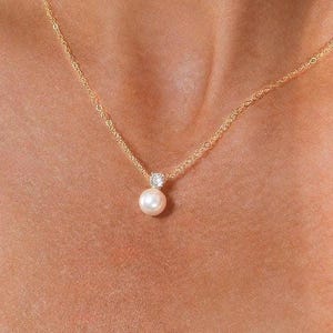 May include: A delicate gold chain necklace featuring a single round white pearl pendant suspended from a small, clear, faceted gemstone setting. The necklace rests against the skin, showcasing the polished luster of the pearl and the gold metal.