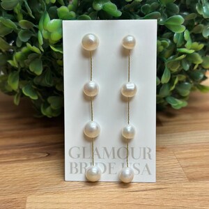 May include: A pair of long, white pearl drop earrings with a gold chain. The earrings are on a white card with the text "Glamour Brides USA".