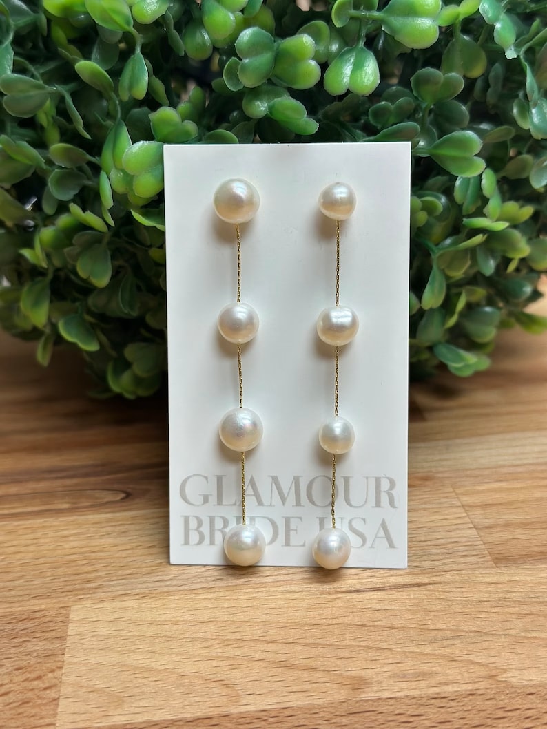 May include: A pair of long, white pearl drop earrings with a gold chain. The earrings are on a white card with the text "Glamour Brides USA".