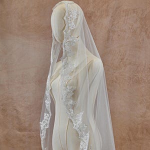 May include: A white bridal veil with delicate lace trim. The veil is draped over a mannequin, showcasing the intricate floral and beaded details along the edges. The sheer fabric creates a soft, ethereal look, ideal for a wedding.