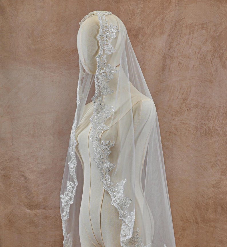 May include: A white bridal veil with delicate lace trim. The veil is draped over a mannequin, showcasing the intricate floral and beaded details along the edges. The sheer fabric creates a soft, ethereal look, ideal for a wedding.