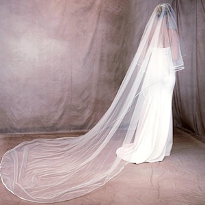 May include: A long, sheer white wedding veil featuring a delicate satin ribbon trim along the edges. The veil is layered, with a shorter top tier and a floor-sweeping train that extends behind the wearer, creating a soft, flowing silhouette.