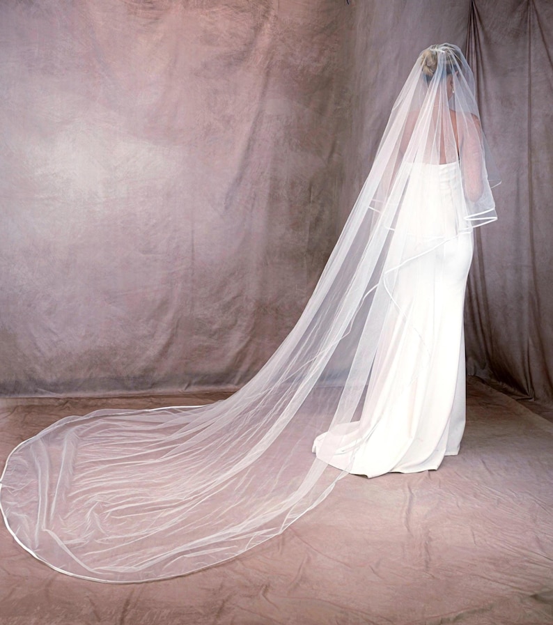 May include: A long, sheer white wedding veil featuring a delicate satin ribbon trim along the edges. The veil is layered, with a shorter top tier and a floor-sweeping train that extends behind the wearer, creating a soft, flowing silhouette.