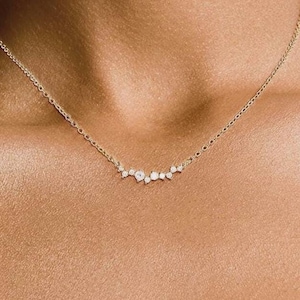 May include: A delicate gold chain necklace featuring a horizontal cluster of small, round, clear white stones arranged in an asymmetrical pattern. The necklace rests against the skin, showcasing a fine metal link chain and a central decorative element.