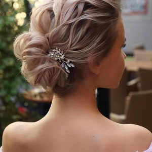 May include: A woman with blonde hair styled in a loose updo with a small jeweled hair clip. The hair is pulled back and styled in a messy bun with loose strands framing the face.