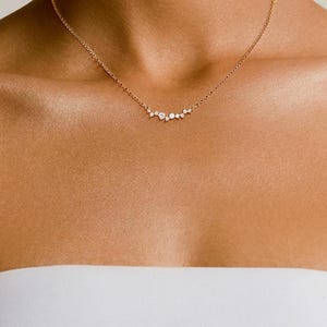 May include: A gold chain necklace featuring a delicate cluster of small, clear, round-cut stones arranged in a horizontal, asymmetrical arc. The stones vary slightly in size, creating a subtle, graduated effect along the center of the chain.