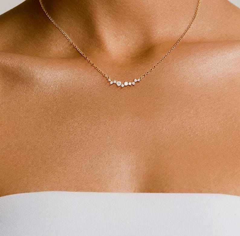 May include: A gold chain necklace featuring a delicate cluster of small, clear, round-cut stones arranged in a horizontal, asymmetrical arc. The stones vary slightly in size, creating a subtle, graduated effect along the center of the chain.