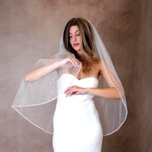 May include: A white bridal veil featuring a subtle shimmering glitter finish and a delicate beaded trim along the edges. The sheer fabric drapes over the shoulders, paired with a strapless white gown that has a clean, fitted bodice design.