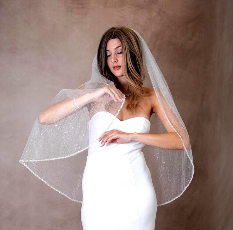 May include: A white bridal veil featuring a subtle shimmering glitter finish and a delicate beaded trim along the edges. The sheer fabric drapes over the shoulders, paired with a strapless white gown that has a clean, fitted bodice design.