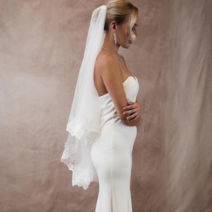 K&ouml;nnte beinhalten: Ein wei&szlig;es Brautkleid mit tr&auml;gerlosem Design und langem, flie&szlig;endem Rock. Ein durchsichtiger, wei&szlig;er Schleier mit Spitzensaum ist am Hinterkopf befestigt. Die Braut tr&auml;gt eine Halskette und Ohrringe.