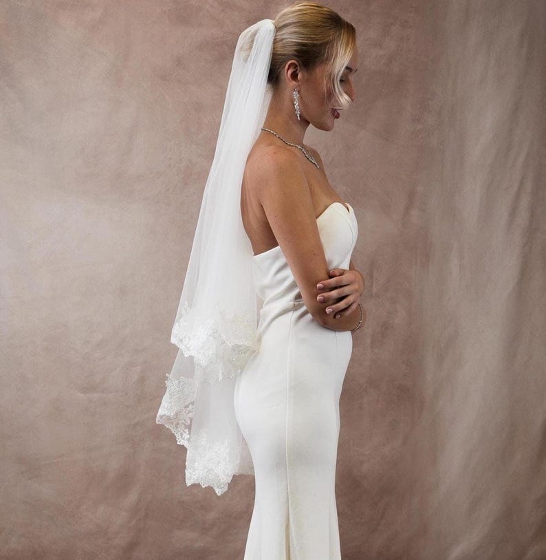 K&ouml;nnte beinhalten: Ein wei&szlig;es Brautkleid mit tr&auml;gerlosem Design und langem, flie&szlig;endem Rock. Ein durchsichtiger, wei&szlig;er Schleier mit Spitzensaum ist am Hinterkopf befestigt. Die Braut tr&auml;gt eine Halskette und Ohrringe.