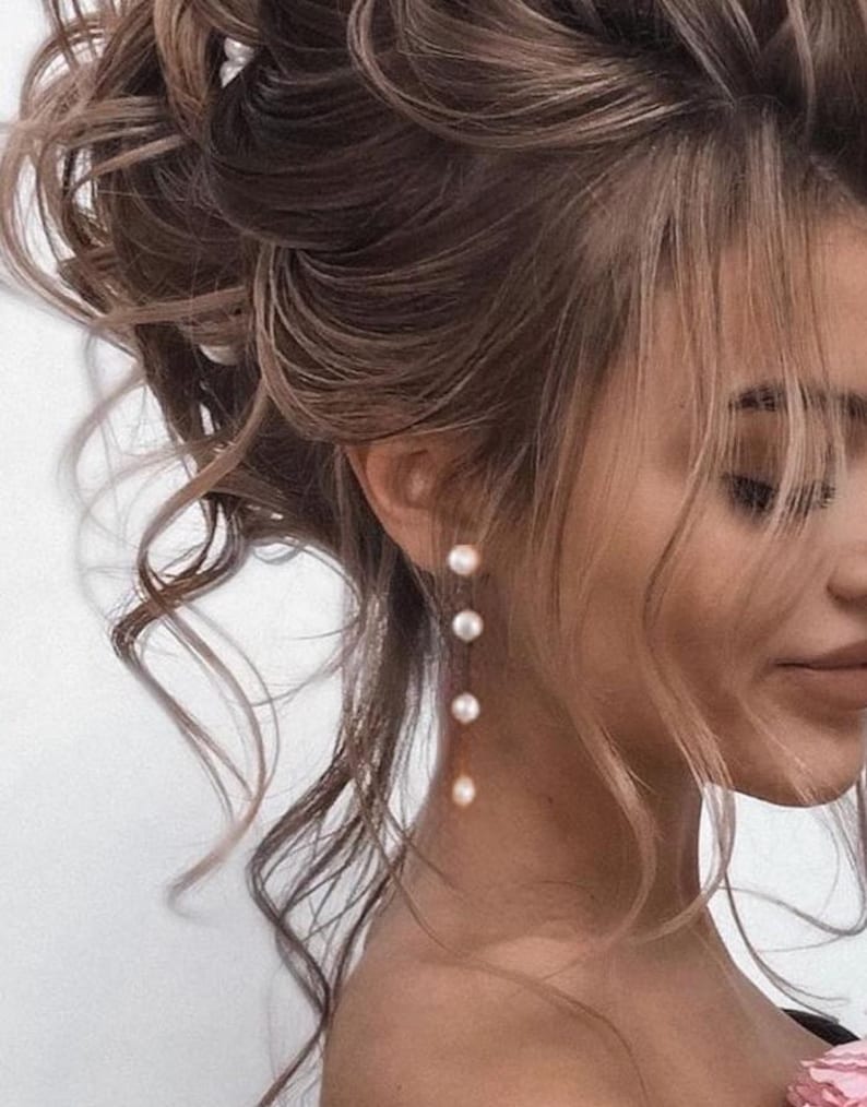 May include: A woman with long, brown, curly hair wearing a pair of pearl drop earrings. The earrings are made of a thin gold chain with five small white pearls.