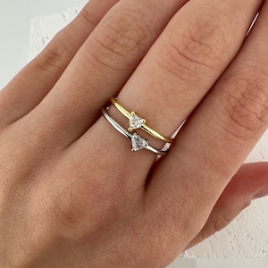 May include: Two delicate rings with heart-shaped diamond accents. The top ring is gold, and the bottom ring is silver.