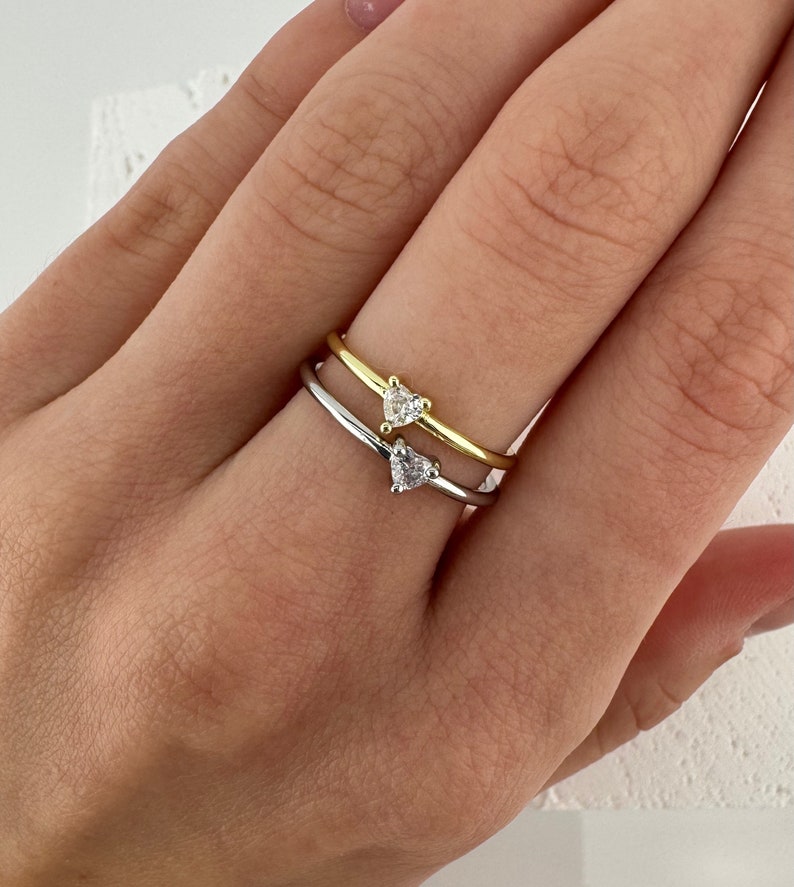 May include: Two delicate rings with heart-shaped diamond accents. The top ring is gold, and the bottom ring is silver.