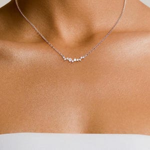 May include: A delicate silver-toned chain necklace featuring a central cluster of small, clear, round-cut stones arranged in an asymmetrical, curved formation that rests along the collarbone, secured with a thin, polished metal link chain.