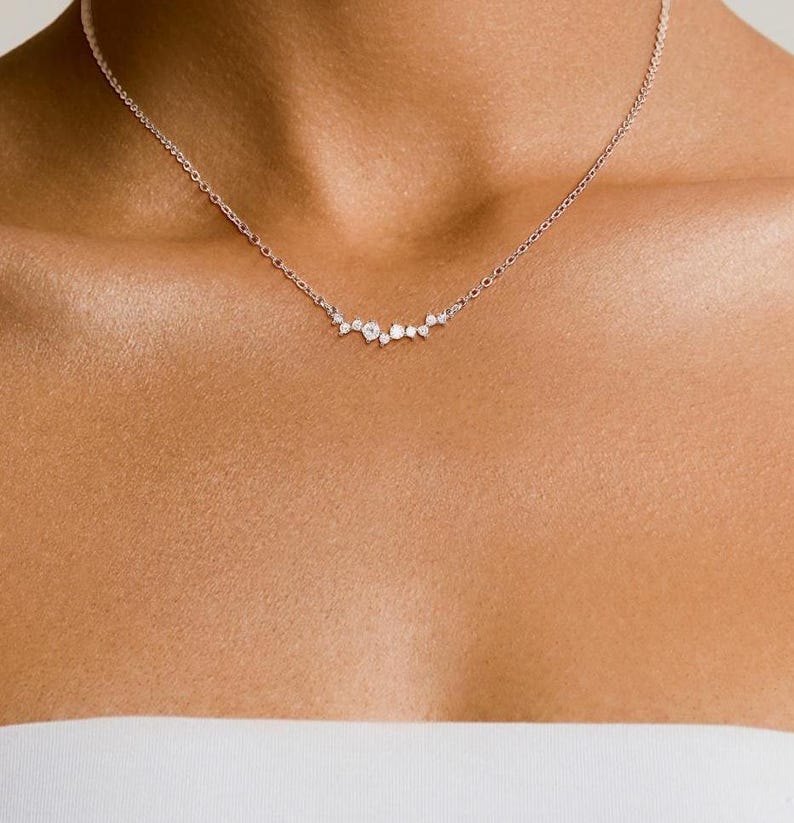 May include: A delicate silver-toned chain necklace featuring a central cluster of small, clear, round-cut stones arranged in an asymmetrical, curved formation that rests along the collarbone, secured with a thin, polished metal link chain.