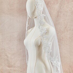 May include: A white bridal veil with delicate floral lace trim is draped over a mannequin. The veil is sheer, allowing the background to be visible. The lace detailing adds a touch of elegance to the wedding accessory.