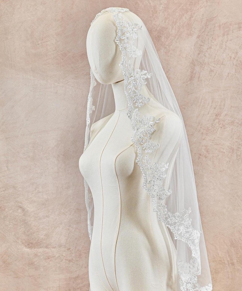 May include: A white bridal veil with delicate floral lace trim is draped over a mannequin. The veil is sheer, allowing the background to be visible. The lace detailing adds a touch of elegance to the wedding accessory.
