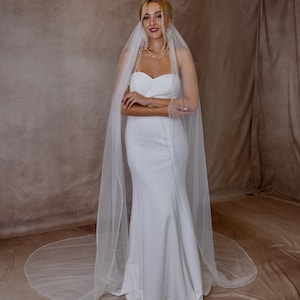 May include: A floor-length white wedding dress with a strapless sweetheart neckline and a fitted bodice that flares into a trumpet skirt. A long, sheer white tulle veil with delicate beaded trim drapes over the shoulders and pools on the floor.