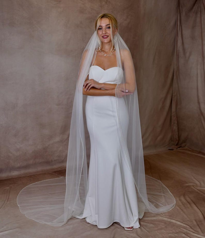 May include: A floor-length white wedding dress with a strapless sweetheart neckline and a fitted bodice that flares into a trumpet skirt. A long, sheer white tulle veil with delicate beaded trim drapes over the shoulders and pools on the floor.