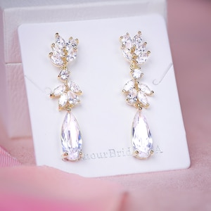 May include: A pair of gold-toned drop earrings with clear, sparkling crystals. The earrings feature a leaf-like design at the top, with a teardrop-shaped crystal pendant. The earrings are displayed on a white card.