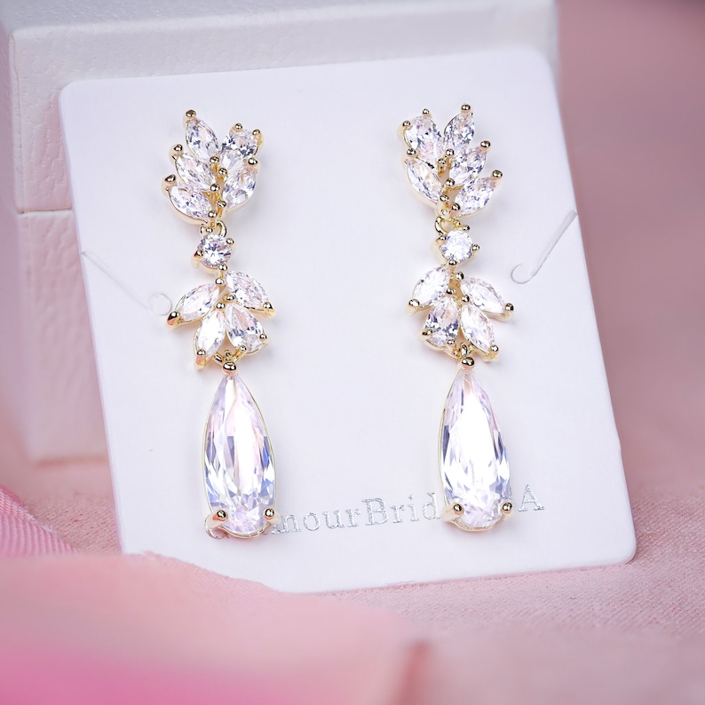 May include: A pair of gold-toned drop earrings with clear, sparkling crystals. The earrings feature a leaf-like design at the top, with a teardrop-shaped crystal pendant. The earrings are displayed on a white card.