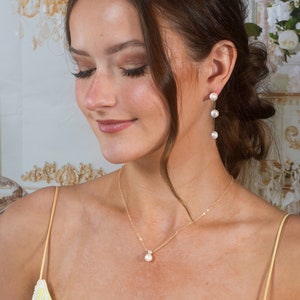 May include: A woman wearing a yellow dress with a delicate gold chain necklace featuring a single pearl pendant. She is also wearing a pair of gold drop earrings with three pearls.