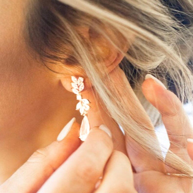 May include: Close-up of a gold-toned earring with a leaf-like design and a teardrop-shaped crystal. The earring is being worn, with the focus on the intricate details of the jewelry. The style is elegant and suitable for special occasions.