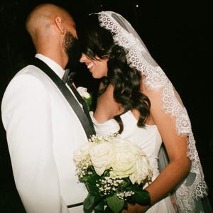 May include: A bride and groom embrace during their wedding ceremony. The bride is wearing a white dress with a lace veil and holds a bouquet of white roses. The groom is wearing a white tuxedo jacket with a black bow tie.