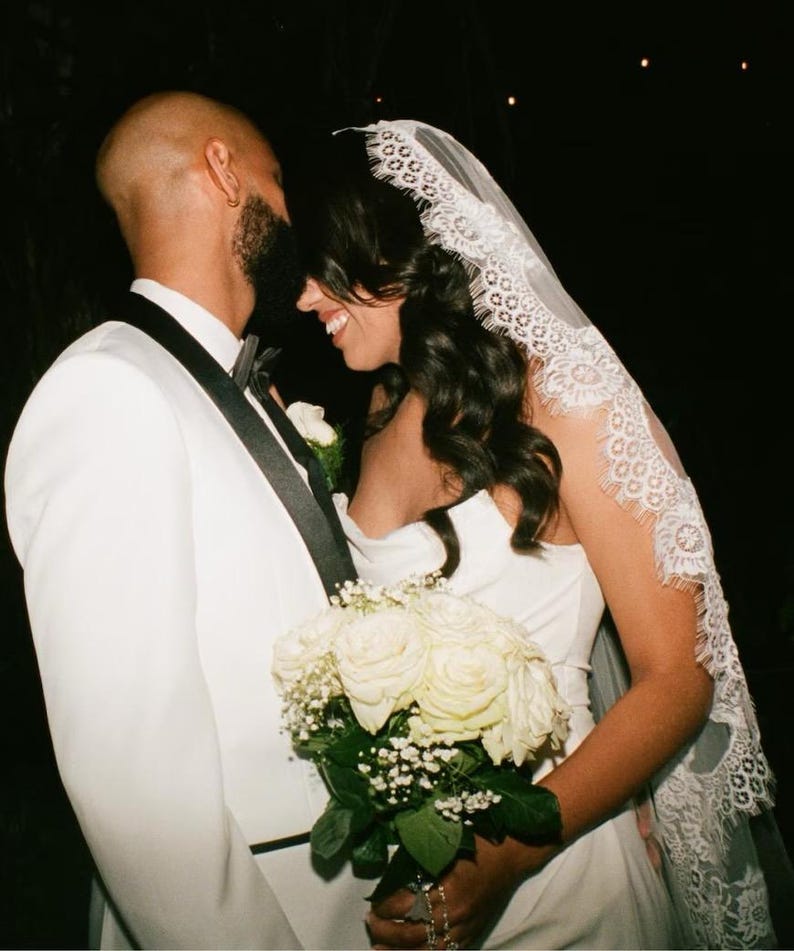 May include: A bride and groom embrace during their wedding ceremony. The bride is wearing a white dress with a lace veil and holds a bouquet of white roses. The groom is wearing a white tuxedo jacket with a black bow tie.