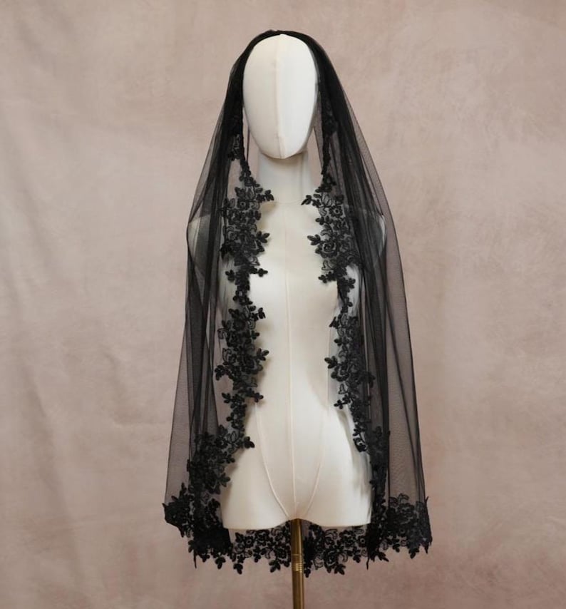 Puede incluir: Un velo negro de tela de tul transparente, con intrincados bordados de encaje floral negro a lo largo de los bordes. El velo est&aacute; colocado sobre un maniqu&iacute;, mostrando el detallado borde de encaje festoneado que enmarca la parte frontal e inferior.