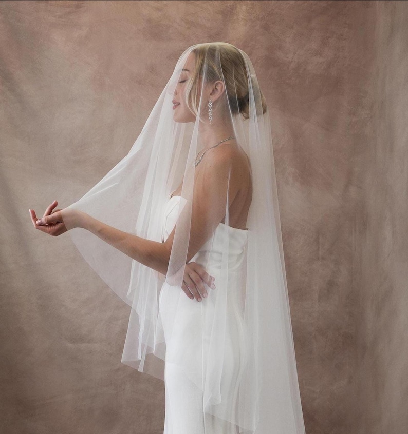 May include: A sheer, floor-length white bridal veil made of fine tulle fabric. The veil is draped over the head and shoulders, extending down the back. It is paired with a strapless white gown, a delicate silver necklace, and drop earrings.