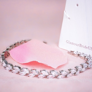 May include: A silver-toned bracelet with clear, marquise-cut stones arranged in a repeating pattern. The bracelet is displayed on a pink surface with a pink fabric and a white card with the text "GlamourBrideUS". The bracelet is a delicate piece of jewelry.