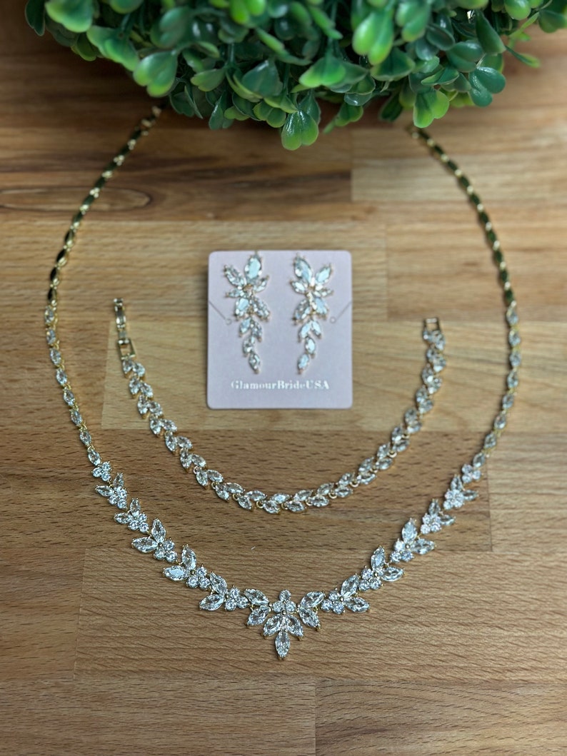 May include: A gold necklace and bracelet set with clear crystal accents. The set also includes a pair of matching earrings. The jewelry is displayed on a wooden surface with a card that reads "GlamourBrideUSA".