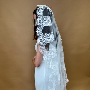 May include: A white bridal veil with a delicate lace trim featuring floral designs. The veil is made of soft tulle and has a scalloped edge.