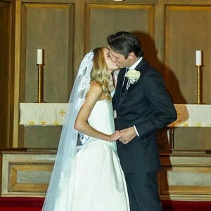 May include: A bride and groom kiss during their wedding ceremony. The bride is wearing a white wedding dress with a lace veil. The groom is wearing a black suit and a white flower.