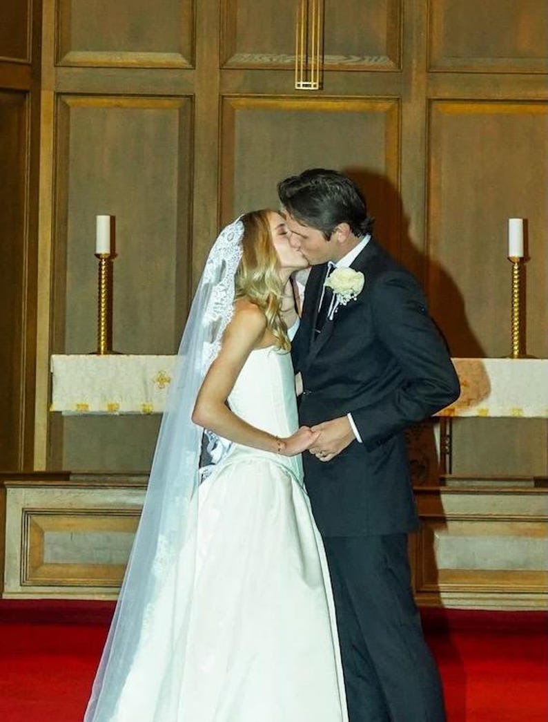 May include: A bride and groom kiss during their wedding ceremony. The bride is wearing a white wedding dress with a lace veil. The groom is wearing a black suit and a white flower.