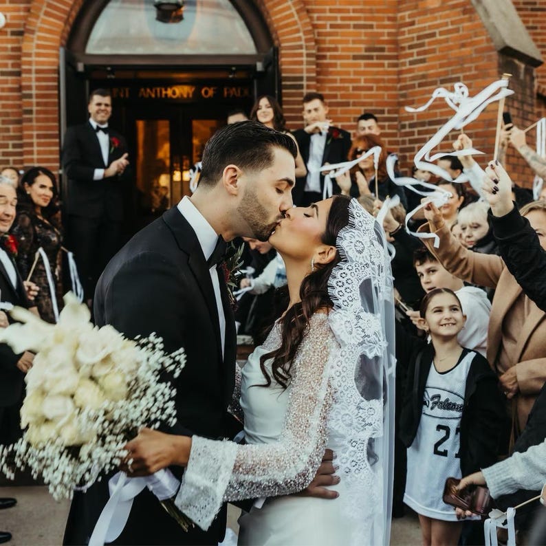 May include: A bride and groom kiss in front of a church, surrounded by guests holding white ribbons. The bride is wearing a white lace dress and veil, and the groom is wearing a black suit. The couple is holding a bouquet of white flowers.