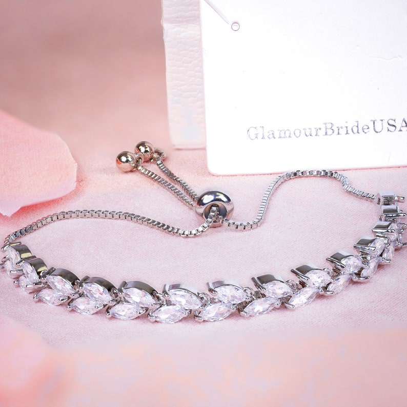May include: A silver-toned bracelet with a leaf-like design, featuring clear, marquise-cut stones. The adjustable bracelet has a sliding clasp and is displayed on a pink surface. The tag reads "GlamourBrideUSA".