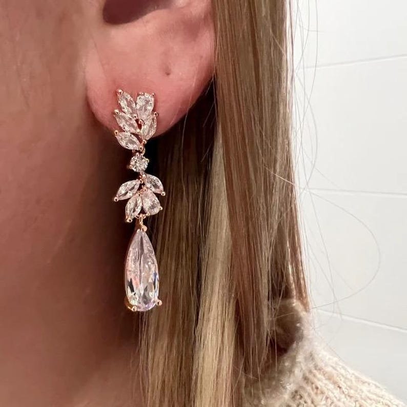 May include: A rose gold earring with a leaf design and a large teardrop-shaped crystal. The earring is worn on the ear of a person with blonde hair. The earring is a dangle style.