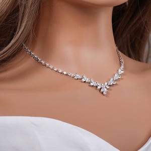 May include: A silver-toned necklace with a delicate design. The necklace features a series of small, clear gemstones set in a floral pattern. The gemstones are arranged in a symmetrical design, creating a sparkling effect. The necklace is designed to sit around the neckline.