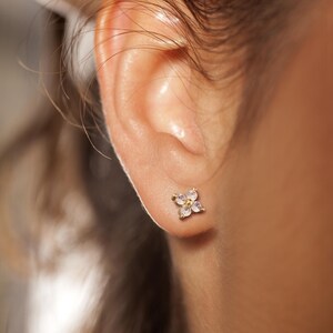 May include: A gold stud earring shaped like a four-petaled flower. The earring features four small, clear, round-cut stones arranged in a cluster, with a tiny gold center point, secured firmly against the earlobe with a metal post backing.